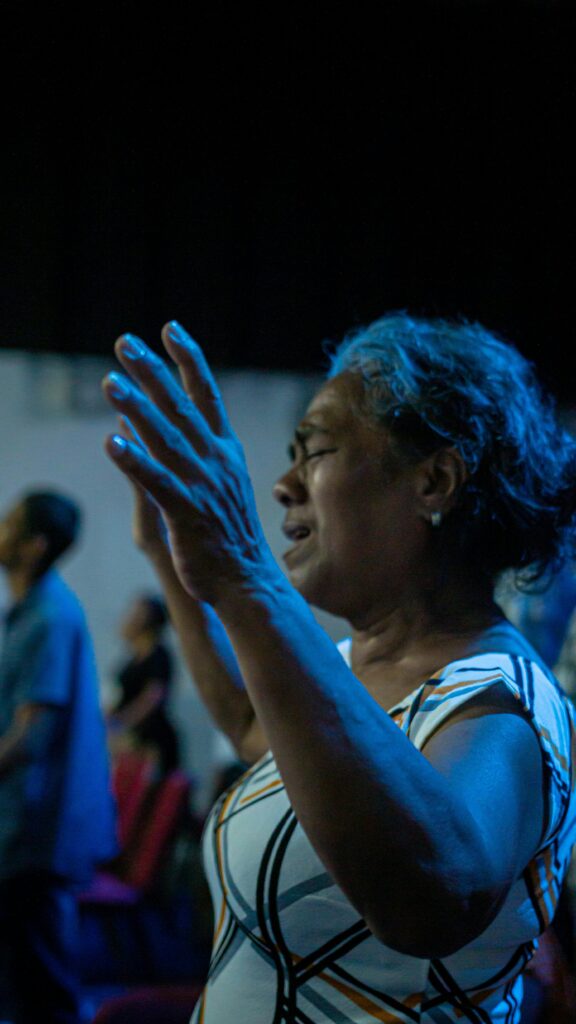 A senior woman praying with raised hands during an indoor religious event, immersed in the spiritual atmosphere.
